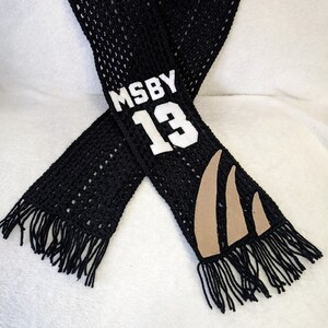 May include: Black crocheted scarf with fringe, featuring a white patch with the text "MSBY 13" and a brown patch with a claw design.