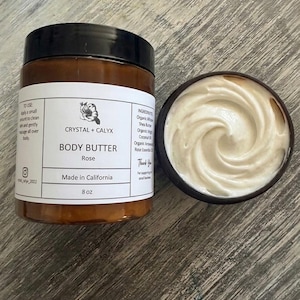 May include: An 8 oz amber plastic jar of Crystal + Calyx Rose body butter with a black lid and a white label featuring text, alongside an open jar revealing a swirled, creamy white substance. The label lists ingredients and usage instructions.