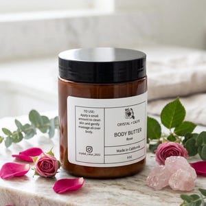May include: An amber-colored plastic jar with a black lid containing body butter. The white label reads Crystal + Calyx Body Butter Rose, Made in California, 8 oz, and includes usage instructions and an Instagram handle, surrounded by rose petals.