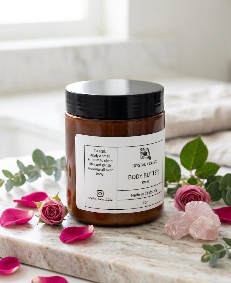 May include: An amber-colored plastic jar with a black lid containing body butter. The white label reads Crystal + Calyx Body Butter Rose, Made in California, 8 oz, and includes usage instructions and an Instagram handle, surrounded by rose petals.