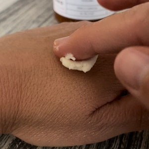 May include: A thick, white, creamy body butter is being applied to the skin. In the background, a blurred amber-colored jar features a white label with black text that reads Body Butter and Lavender, indicating the product's scent and category.