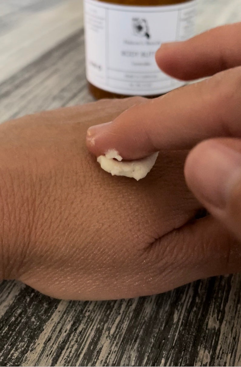 May include: A thick, white, creamy body butter is being applied to the skin. In the background, a blurred amber-colored jar features a white label with black text that reads Body Butter and Lavender, indicating the product's scent and category.