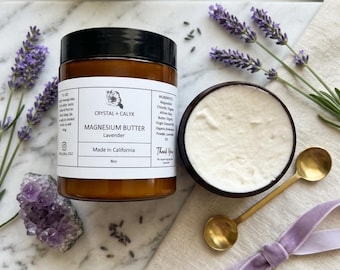 Op de afbeelding: Een amberkleurige pot Crystal + Calyx Magnesium Butter Lavender met een zwarte deksel, gelabeld 8oz, naast een open pot met witte crème, een gouden lepel, een paars amethistkristal, lavendeltakjes en een lichtpaars fluwelen lint.