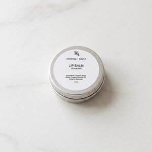May include: A round silver metal tin containing unscented lip balm. The white label features the text CRYSTAL + CALYX, LIP BALM, Unscented, and lists ingredients including Organic Shea Butter, Organic Almond Oil, and Organic Beeswax, with a net weight of 0.5oz.