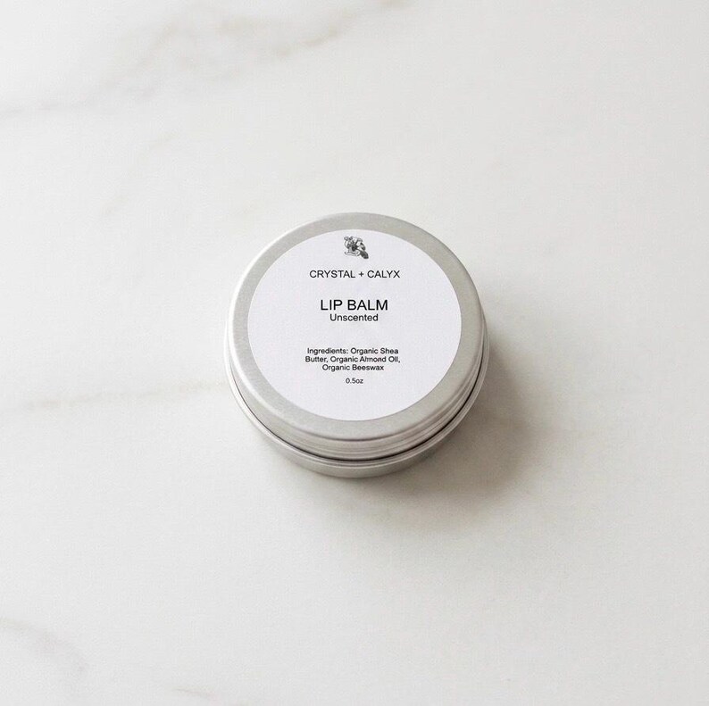 May include: A round silver metal tin containing unscented lip balm. The white label features the text CRYSTAL + CALYX, LIP BALM, Unscented, and lists ingredients including Organic Shea Butter, Organic Almond Oil, and Organic Beeswax, with a net weight of 0.5oz.