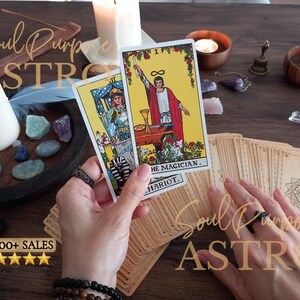 May include: Two tarot cards, The Magician and The Chariot, are held up. The Magician card features a figure in a red robe on a yellow background. The Chariot card shows a figure in blue armor. A deck of cards with a geometric back design lies below.