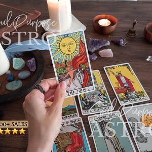May include: A hand holds The Sun tarot card featuring a bright yellow sun, a child on a white horse, and sunflowers. Other tarot cards including The Magician, The Chariot, and The Hermit are spread out alongside candles, crystals, and a small bell.
