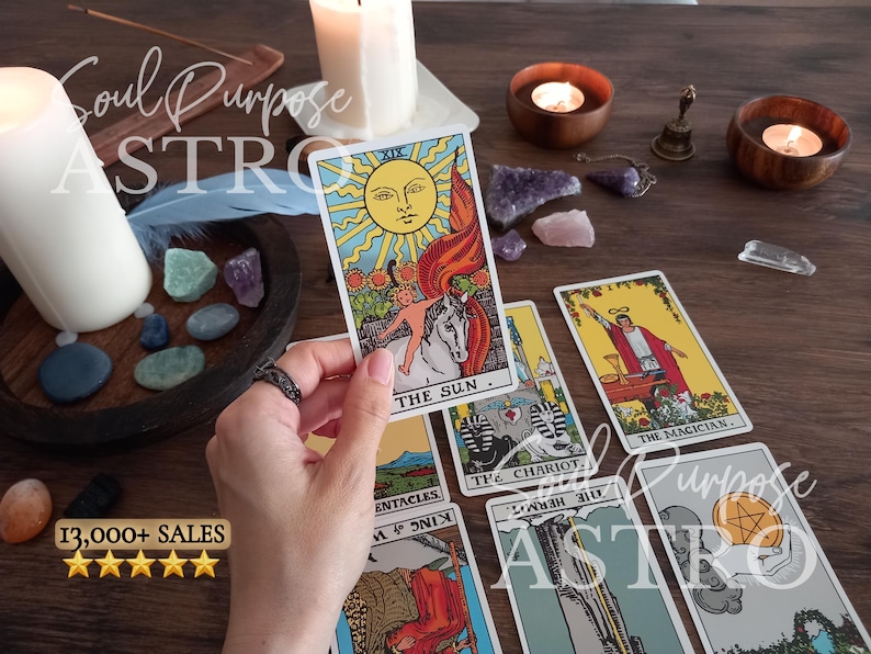May include: A hand holds The Sun tarot card featuring a bright yellow sun, a child on a white horse, and sunflowers. Other tarot cards including The Magician, The Chariot, and The Hermit are spread out alongside candles, crystals, and a small bell.