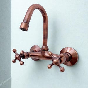 Puede incluir: Un grifo de pared de color cobre con un caño curvo y dos manijas en forma de cruz. El grifo es de metal y tiene una apariencia vintage. El accesorio está montado en una pared azul claro.