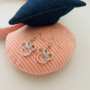 May include: Silver-toned dangle earrings in the shape of a mouse head with a bow, each adorned with clear crystals. The earrings are displayed on a pink corduroy surface, with a denim blue bow in the background.