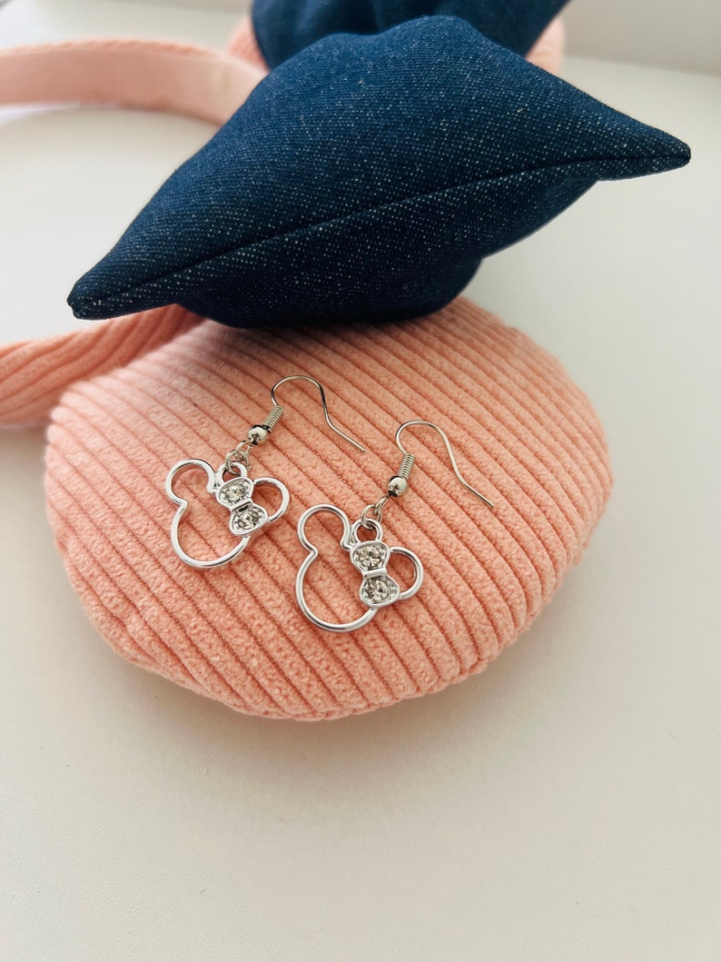 May include: Silver-toned dangle earrings in the shape of a mouse head with a bow, each adorned with clear crystals. The earrings are displayed on a pink corduroy surface, with a denim blue bow in the background.