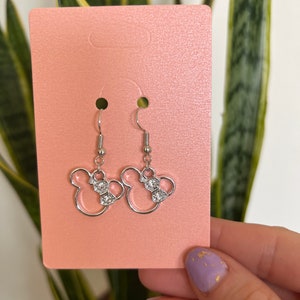 May include: Silver-toned dangle earrings featuring a Minnie Mouse design. Each earring has a bow with clear, sparkling accents. The earrings are displayed on a pink card, with a green plant in the background.