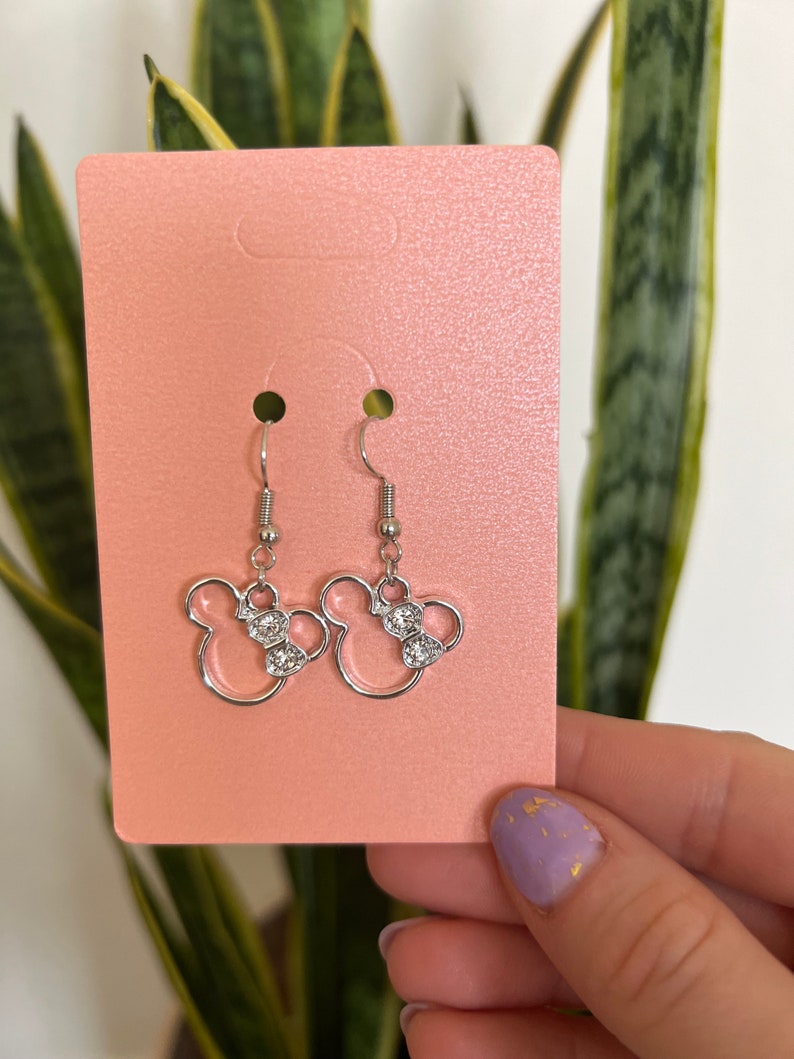 May include: Silver-toned dangle earrings featuring a Minnie Mouse design. Each earring has a bow with clear, sparkling accents. The earrings are displayed on a pink card, with a green plant in the background.