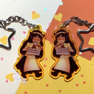 May include: Two keychains featuring a cartoon character with brown hair, wearing a white shirt and a black apron, holding a tray of pastries. The character is depicted with a surprised expression. The keychains are made of clear acrylic and have a silver star-shaped charm attached.