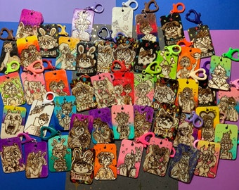Puede incluir: Una colección de llaveros de madera coloridos con varios personajes de dibujos animados y diseños. Los llaveros están decorados con colores vibrantes y detalles intrincados, incluyendo un conejo, un gato y una variedad de otras figuras caprichosas. Los llaveros están unidos a anillos de metal y están listos para ser usados.