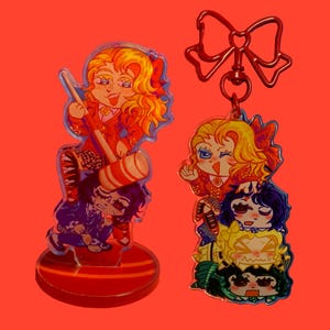 May include: Two acrylic charms with cartoon illustrations. One charm features a character with orange hair, a red outfit, and a striped mallet. The other charm has a bow-shaped clasp and depicts three stacked characters.