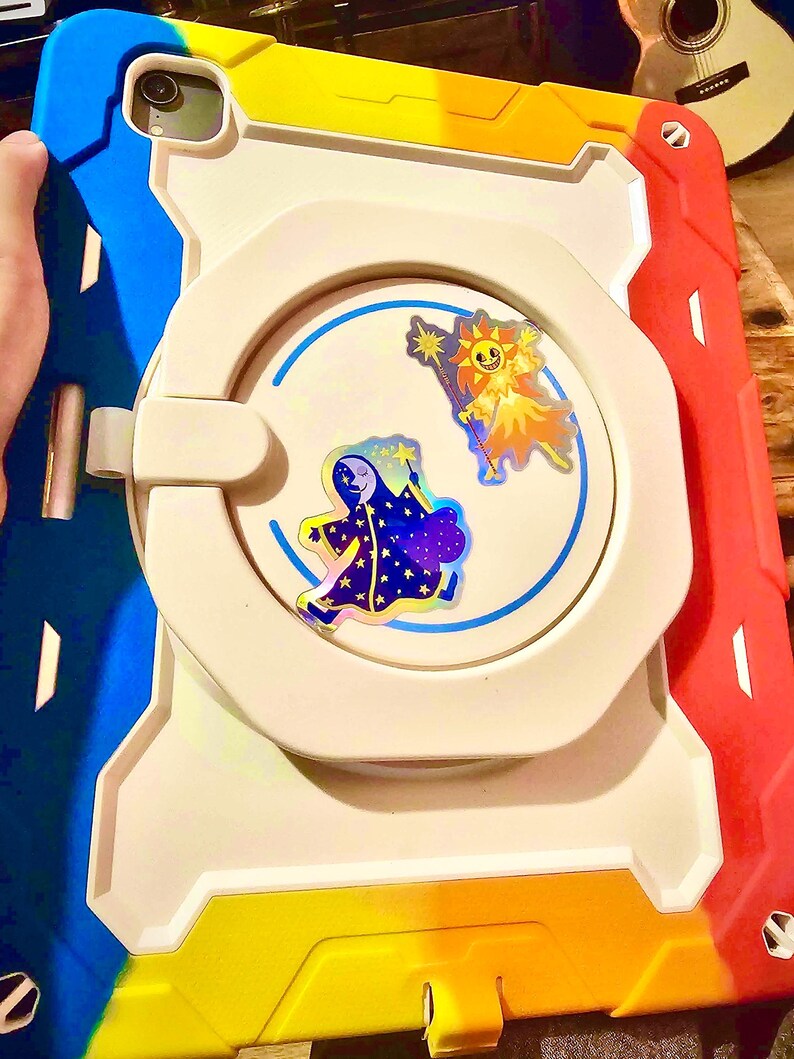 May include: A multi-colored tablet case with a white center and a blue, yellow, and red border. The case features two cartoon stickers of a sun and a moon. The case is designed to protect a tablet.