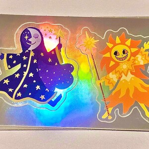 May include: Holographic stickers featuring whimsical illustrations of the moon and sun. The moon character wears a starry robe and holds a star wand. The sun character is orange and yellow, holding a sun wand. The stickers have a shiny, iridescent finish.