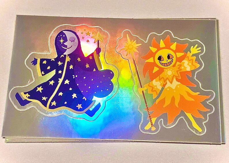 May include: Holographic stickers featuring whimsical illustrations of the moon and sun. The moon character wears a starry robe and holds a star wand. The sun character is orange and yellow, holding a sun wand. The stickers have a shiny, iridescent finish.