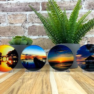 May include: Four round coasters with sunset scenes. Each coaster features a different image of a sunset over water, with docks, bridges, and trees silhouetted against the colorful sky. They are displayed on clear acrylic stands.