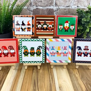 May include: A collection of square decorative tiles featuring gnome illustrations. Each tile has a different holiday or seasonal theme, including Halloween, Christmas, and St. Patrick's Day. The tiles are displayed on small wooden easels.