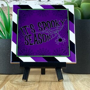 May include: A square Halloween-themed decorative tile with the text "IT'S SPOOKY SEASON" in white, surrounded by a purple background with spiderwebs. The tile has a black, white, and purple striped border and is displayed on a small black easel.