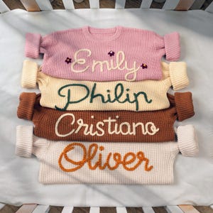 May include: Four knitted sweaters stacked on top of each other. The sweaters are in different colors and have names embroidered on them: Emily, Philip, Cristiano, and Oliver.