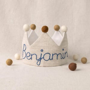 May include: A linen crown with the name "Benjamin" embroidered in blue. The crown is decorated with brown and white pom-poms. Several small felt balls in matching colors are scattered around the crown.