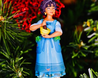 May include: A Christmas tree ornament featuring a woman in a blue dress holding a mortar and pestle. The ornament has a green base and is hanging from a blue ribbon. The background is a Christmas tree with red and green accents.