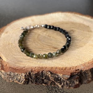 May include: A beaded bracelet featuring faceted green and black gemstones. The bracelet is strung on a silver chain and clasp. The jewelry is displayed on a natural wood slice with a rough bark edge.