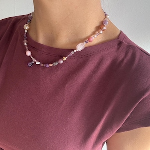 Peut inclure: Un collier de perles composé de pierres précieuses roses, violettes et transparentes. Le collier, orné d'un pendentif, est porté sur un t-shirt bordeaux.
