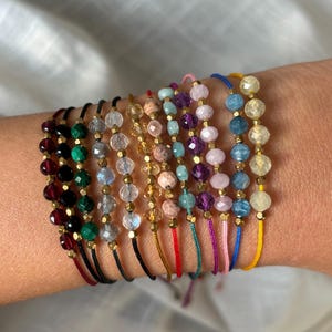 Puede incluir: Una pila de pulseras coloridas con diferentes cuentas de piedras preciosas y detalles dorados. Cada pulsera tiene un cordón de color diferente.