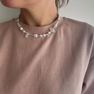 Könnte beinhalten: Eine Halskette mit weißen Perlen, klaren Perlen und unregelmäßig geformten Steinen. Die Halskette wird von einer Person getragen, die ein mauvefarbenes Rundhals-Oberteil trägt.