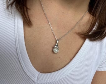 May include: A silver necklace with a pendant featuring a white pearl. The pendant is held by a decorative silver setting. The necklace is worn against a white t-shirt.