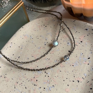 May include: A delicate necklace featuring a dark, beaded chain with two iridescent, round gemstones. The necklace is displayed on a speckled, light-colored surface, with a blurred background including a candle and a decorative box.