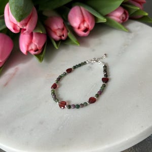 Op de afbeelding: Een zilveren armband met groene en rode edelsteen kralen. De kralen zijn in de vorm van harten. De armband ligt op een wit marmeren oppervlak.