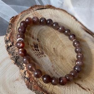 Puede incluir: Un brazalete de cuentas de piedra de sol marrón con una cuenta espaciadora de plata. El brazalete está sobre una superficie de madera.