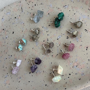 Op de afbeelding: Een collectie oorknopjes van ruwe edelstenen met diverse mineralen zoals groene malachiet, metallic pyriet, roze toermalijn, paarse amethist, lichtblauwe fluoriet, turkoois en iriserende opaal, allemaal bevestigd op zilveren stekers.