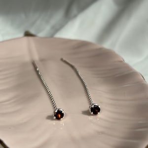 May include: A pair of silver threader earrings with a small, round, dark red gemstone at the end of each chain. The earrings are displayed on a light pink, leaf-shaped dish.