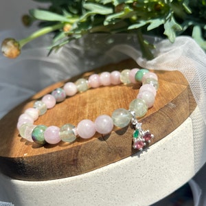 May include: A pink and green gemstone bracelet with a silver charm in the shape of a cherry. The charm has small red and green gemstones.