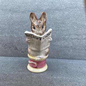 May include: A ceramic figurine of a brown rabbit reading a newspaper titled "The Tailor Cutter." The rabbit is perched atop a spool of thread with pink and yellow accents. A charming decorative piece.