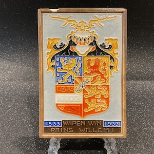 May include: A ceramic tile depicting a coat of arms with a lion and a crown. The tile is decorated with gold, red, blue and white paint. The text on the tile reads "1533 Wapen Van 1933 Prins Willem I".