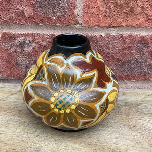 Può includere: Un piccolo vaso in ceramica dipinto a mano con un motivo floreale marrone, giallo e bianco. Il vaso ha un corpo arrotondato e un bordo nero.
