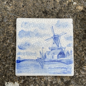 Può includere: Una piastrella di ceramica blu e bianca con la raffigurazione di un mulino a vento e una scena di un villaggio olandese tradizionale.