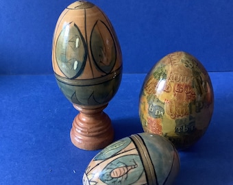 May include: Three decorative wooden eggs, each with a different painted design. The first egg is brown with green and gold accents, the second egg is yellow with brown, green, and red accents, and the third egg is blue with green and gold accents.
