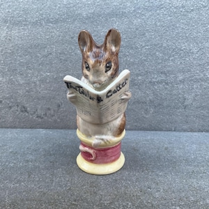Puede incluir: Figura de cerámica de un conejo marrón y blanco leyendo un periódico titulado "The Tailor & Cutter". El conejo está posado sobre una bobina de hilo con una banda rosa y una base amarilla. Estética vintage.
