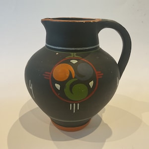 Könnte beinhalten: Schwarzer Keramik Krug mit einem bemalten Design aus drei stilisierten Wirbeln in Orange, Grün, Blau und Weiß. Der Krug hat einen Henkel und einen runden Körper.