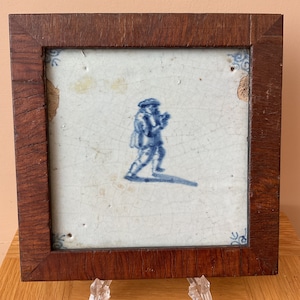 May include: A blue and white Delft tile depicting a man in a hat and coat walking. The tile is framed in a dark wood frame.