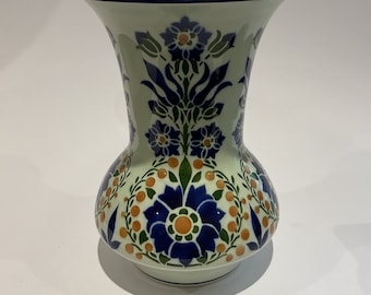 May include: A white ceramic vase with a blue and green floral pattern. The vase has a wide mouth and a rounded base.
