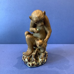 May include: A brown ceramic figurine of a monkey holding a smaller monkey. The larger monkey is sitting on a textured base with brown and white accents.